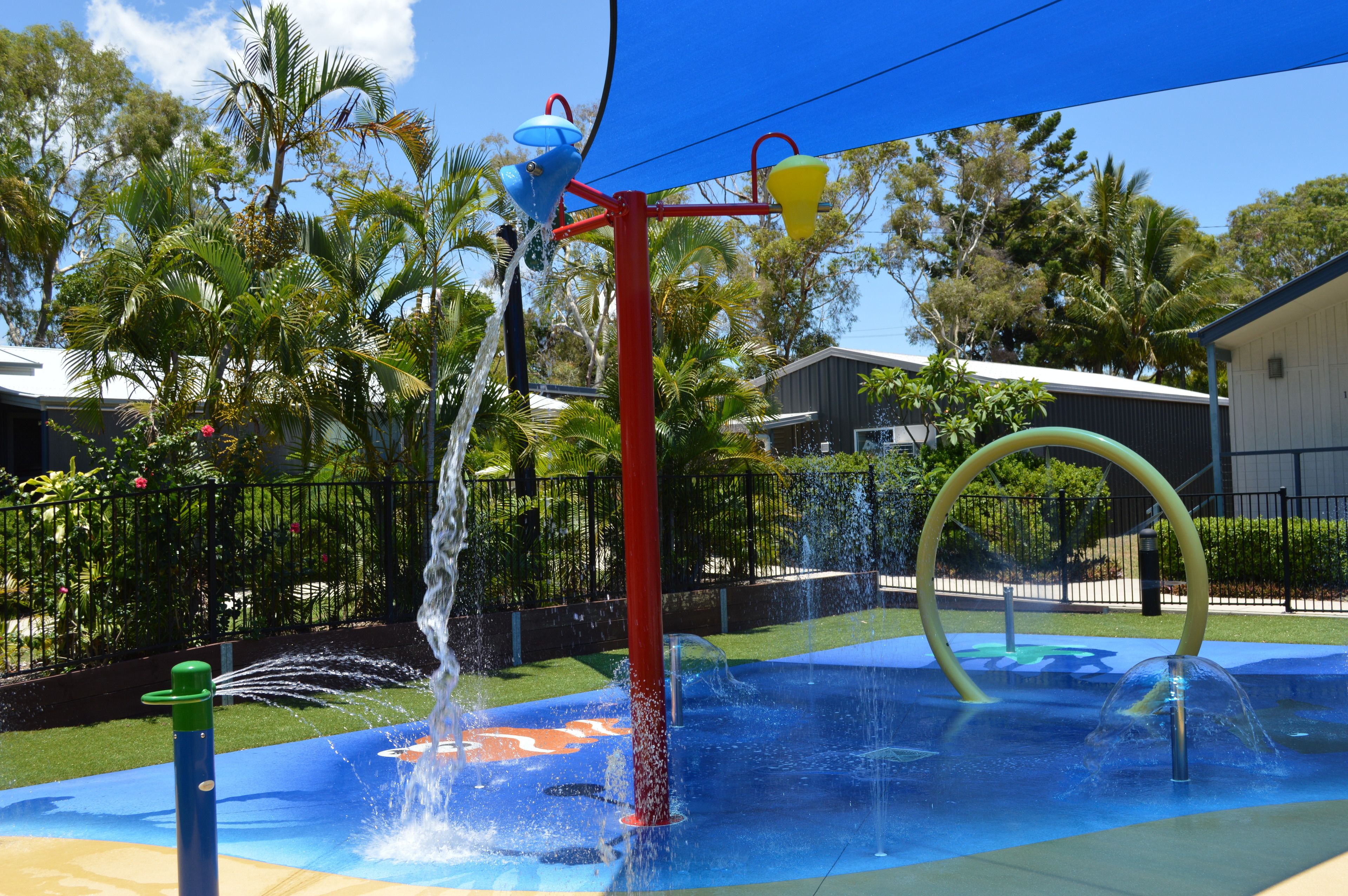 A water play area with multiple water features, including a fountain and water jets, surrounded by palm trees and a fenced area.