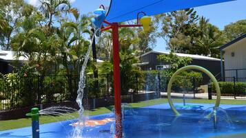 A water play area with multiple water features, including a fountain and water jets, surrounded by palm trees and a fenced area.