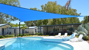 Piscina con sillones de descanso y zona sombreada, rodeada de edificios y árboles.