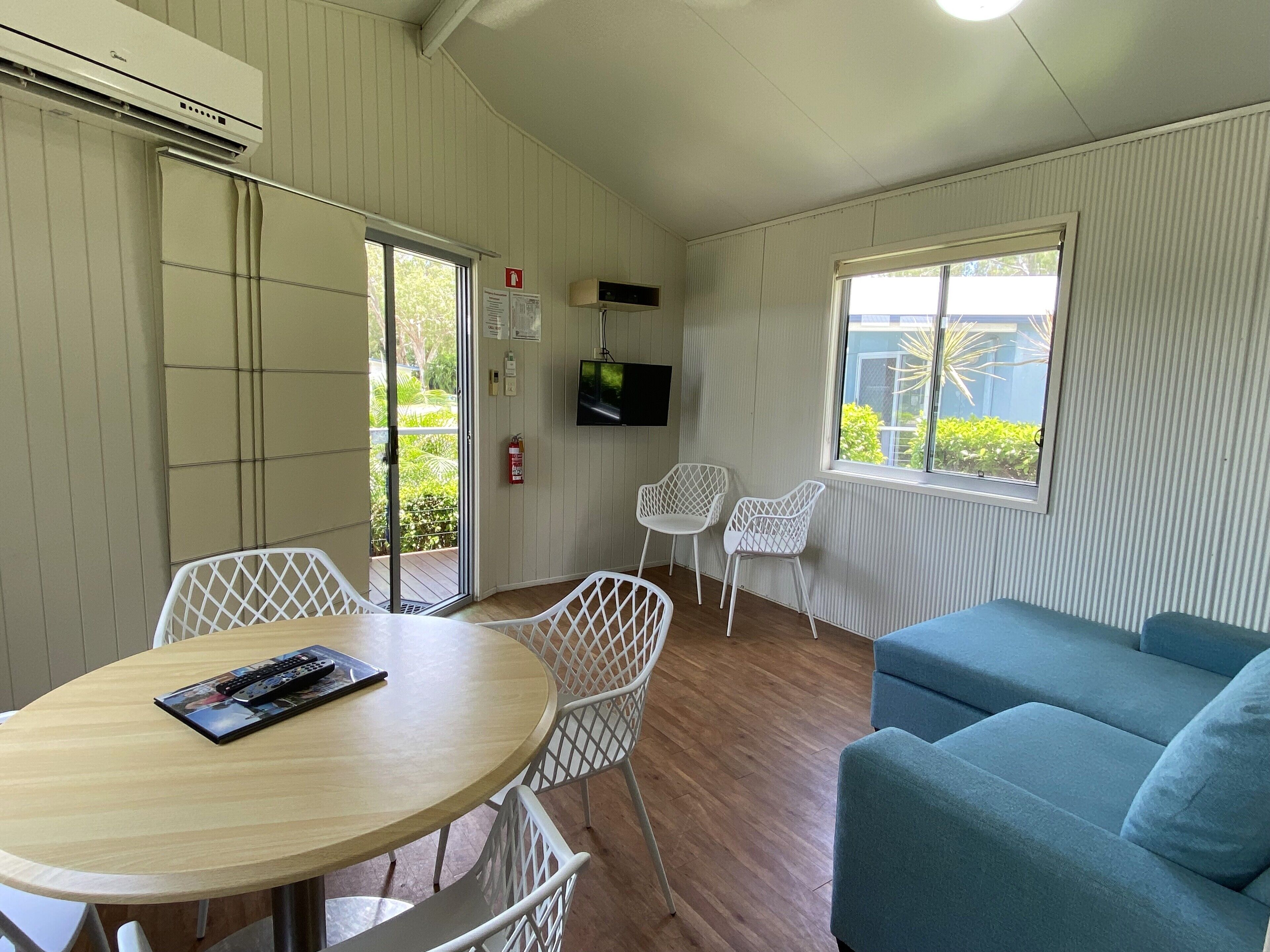 A room with a round wooden table, white chairs, a blue sofa, a TV mounted on the wall, and a window with a view of greenery.