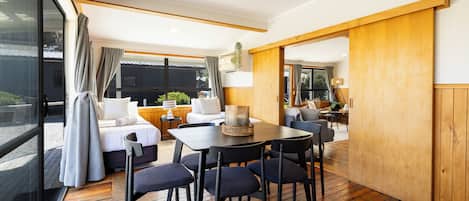 Quarto moderno e bem iluminado, com piso de madeira, mesa de jantar com cadeiras, sofá e uma cama com cortinas.