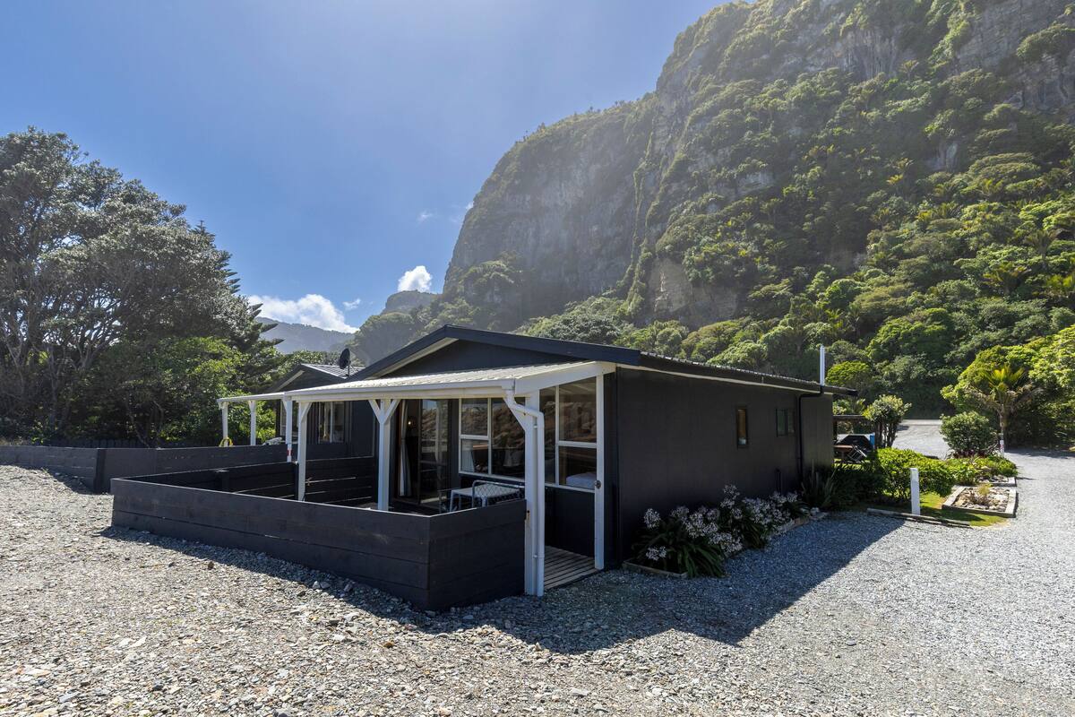 Um edifício pequeno e moderno com um alpendre coberto, rodeado por um caminho de cascalho e vegetação exuberante.