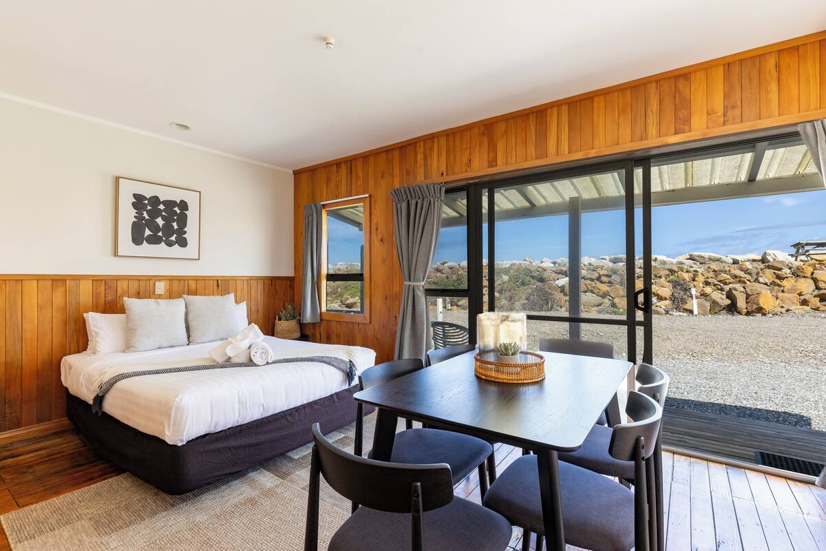 Quarto com cama, mesa, cadeiras, janela com cortinas e vista para rochas e céu.
