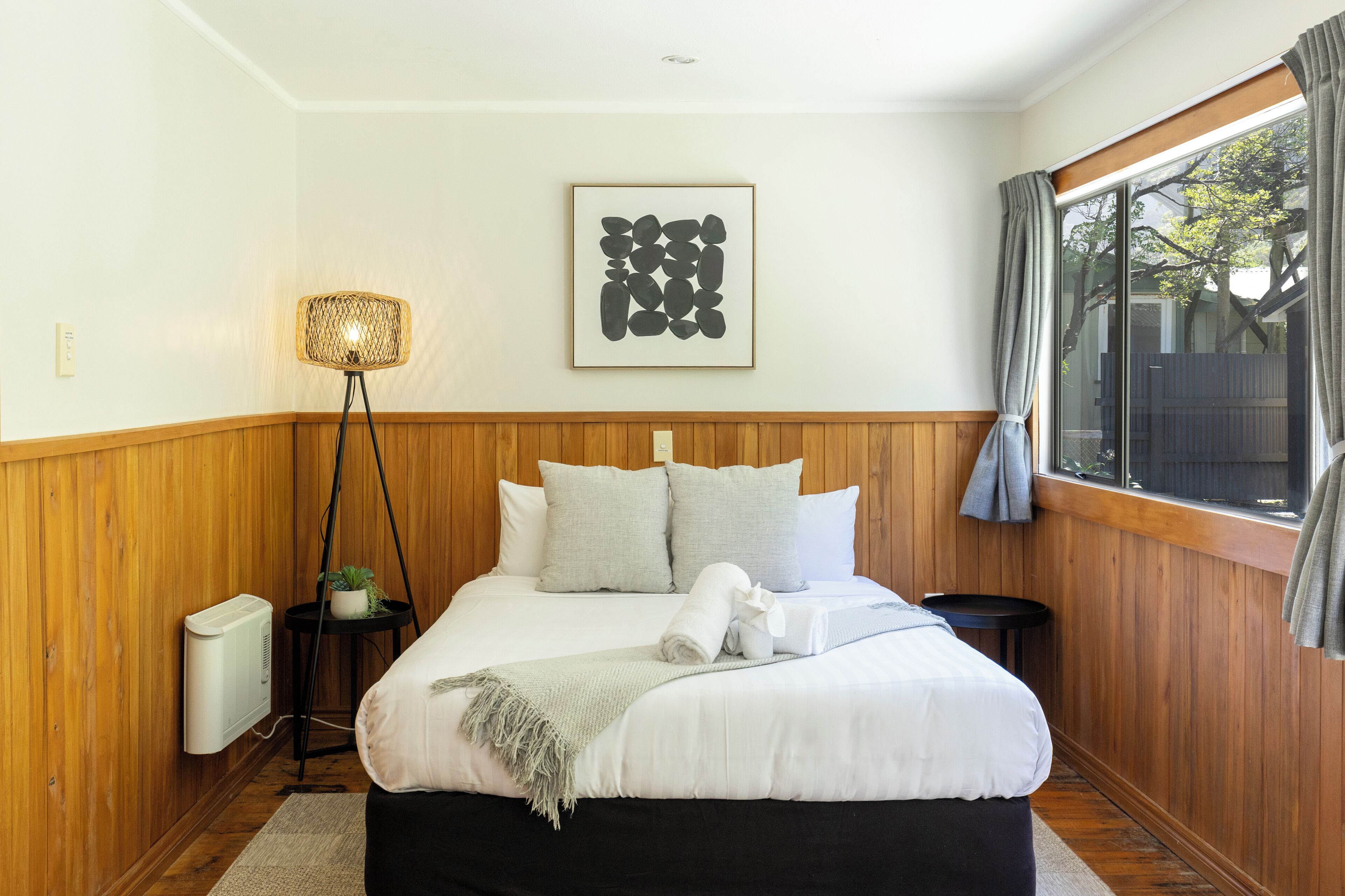 A bedroom with a wooden paneled wall, a bed with white bedding, a framed abstract artwork, a standing floor lamp, and a window with curtains.