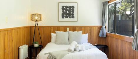 A bedroom with a wooden paneled wall, a bed with white bedding, a framed abstract artwork, a standing floor lamp, and a window with curtains.