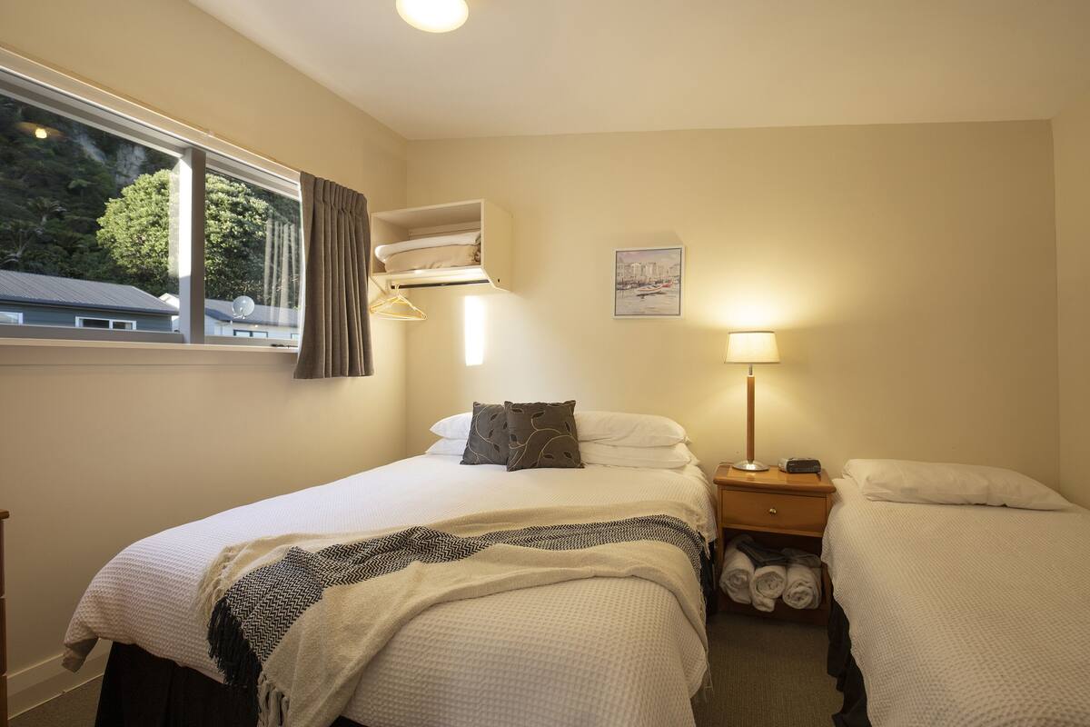Quarto com duas camas, uma janela com cortinas, luminária de cabeceira e prateleira na parede com toalhas.