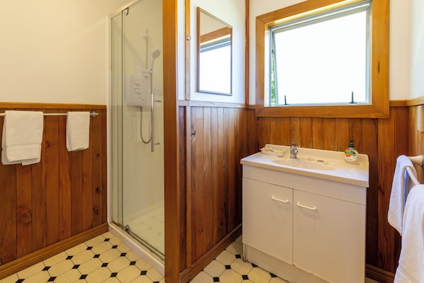 A bathroom with a glass shower enclosure, wooden paneling, a white sink with a mirror above it, and a window providing natural light.