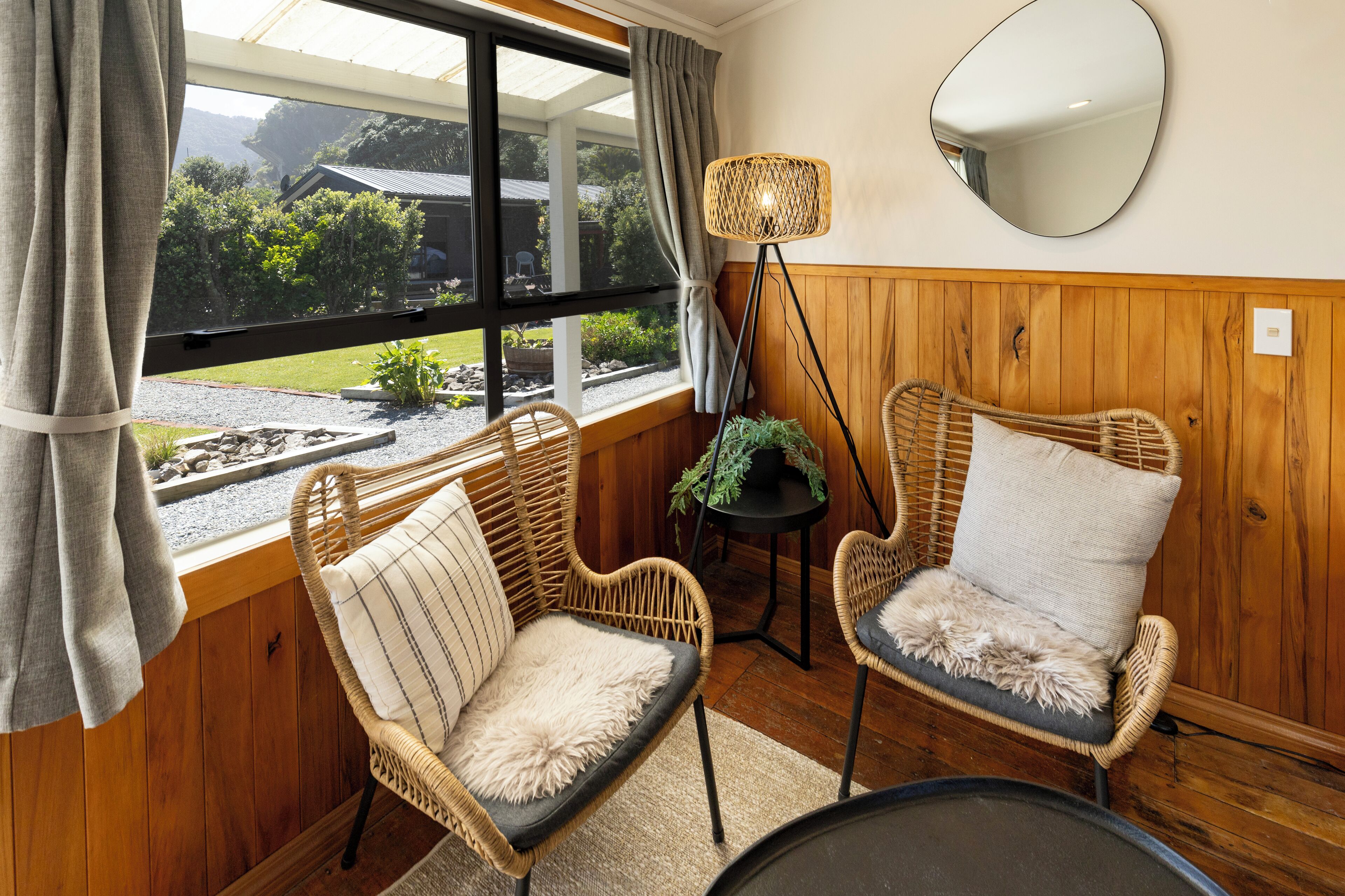 A cozy room with wicker chairs, a round mirror, and a view of greenery outside.