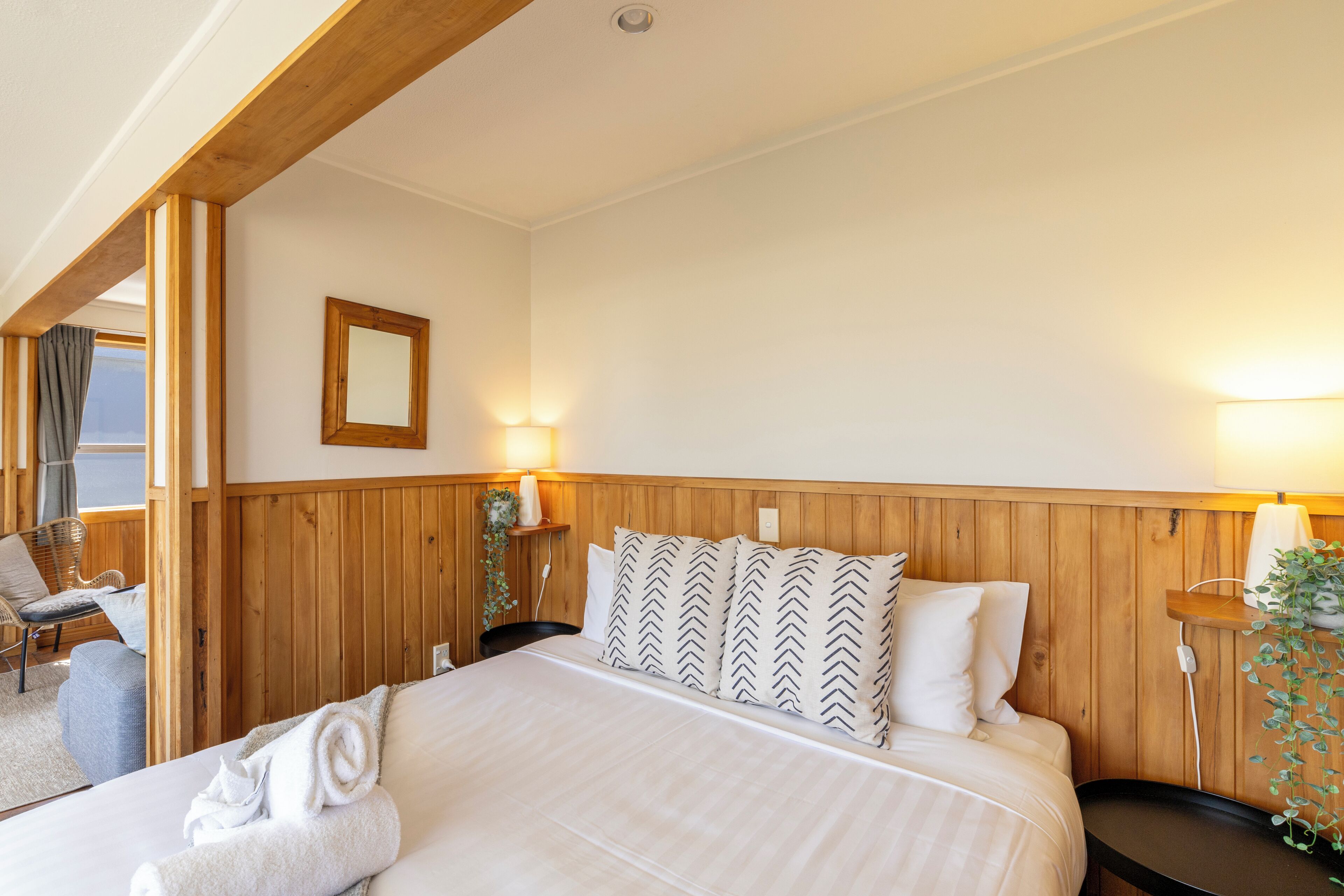 Una habitación de hotel con cabecera de madera, cama con ropa de cama blanca, dos lámparas de noche, una silla y una ventana con cortinas.