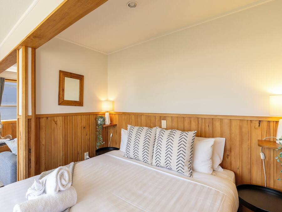 Una habitación de hotel con cabecera de madera, cama con ropa de cama blanca, dos lámparas de noche, una silla y una ventana con cortinas.