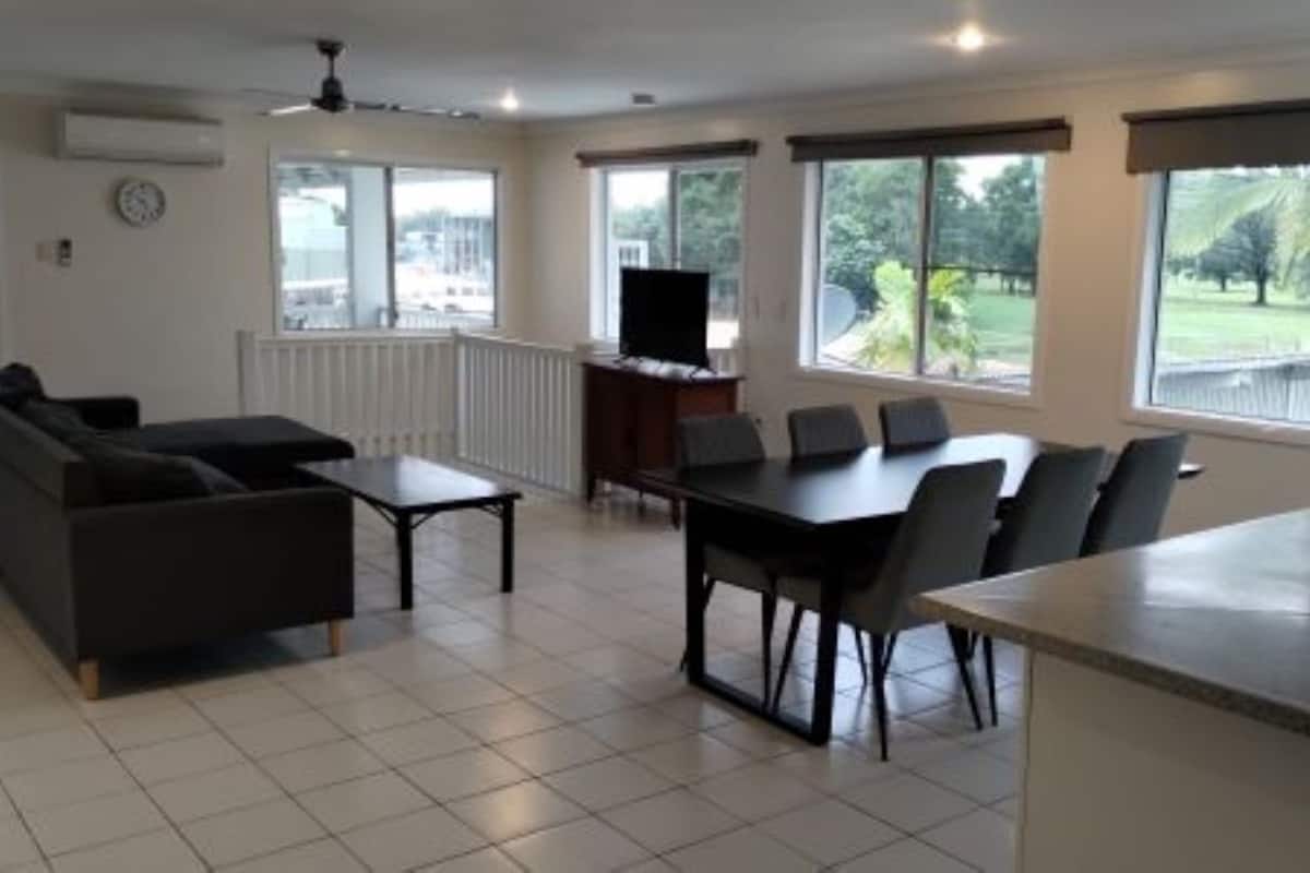 A spacious living area with a sofa, a dining table with chairs, a TV, and large windows offering a view of the outdoors.