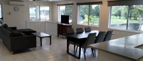 A spacious living area with a sofa, a dining table with chairs, a TV, and large windows offering a view of the outdoors.
