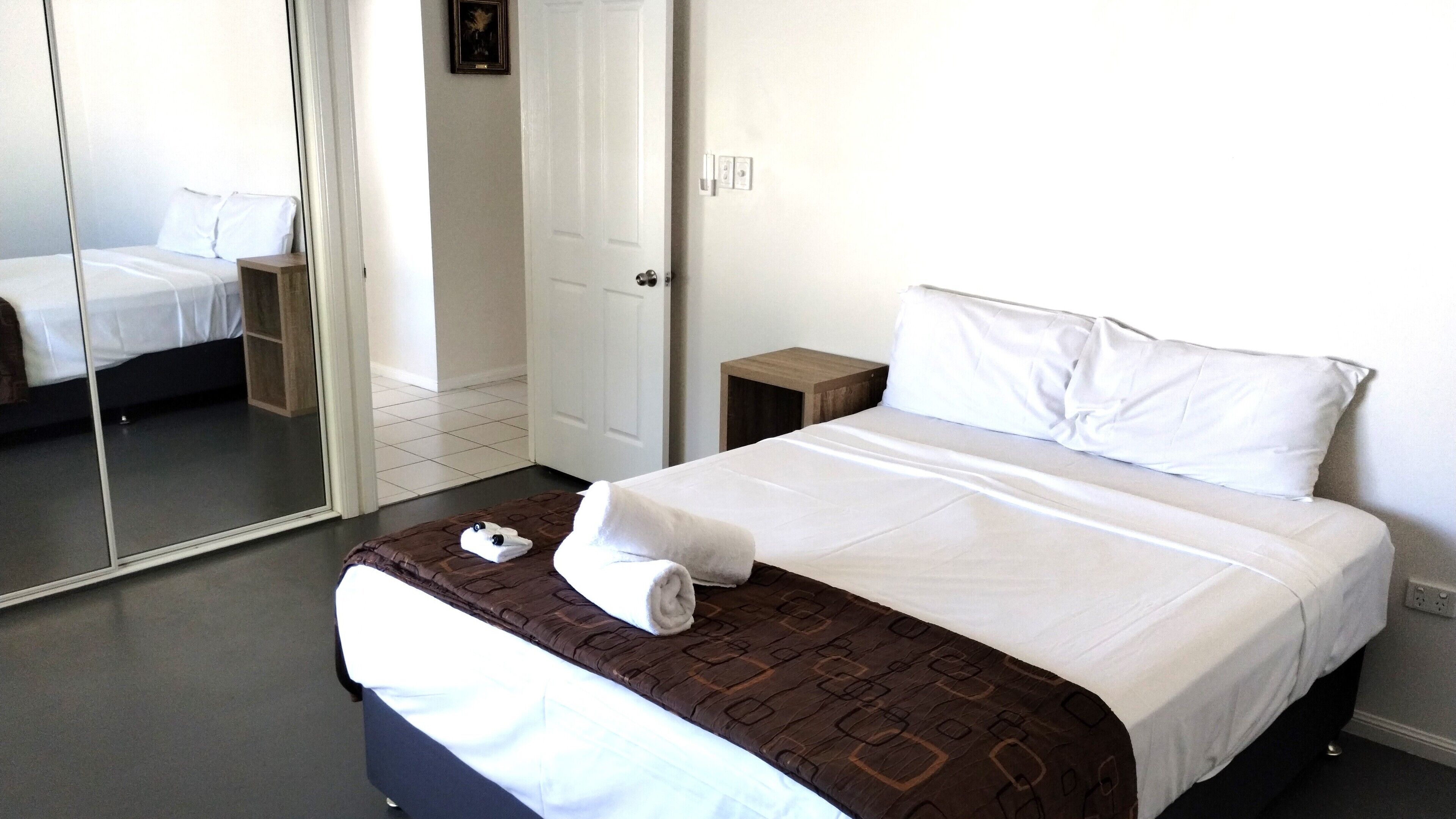A hotel room with a large mirror, a bed with white linens, a brown patterned throw, and a small wooden nightstand.
