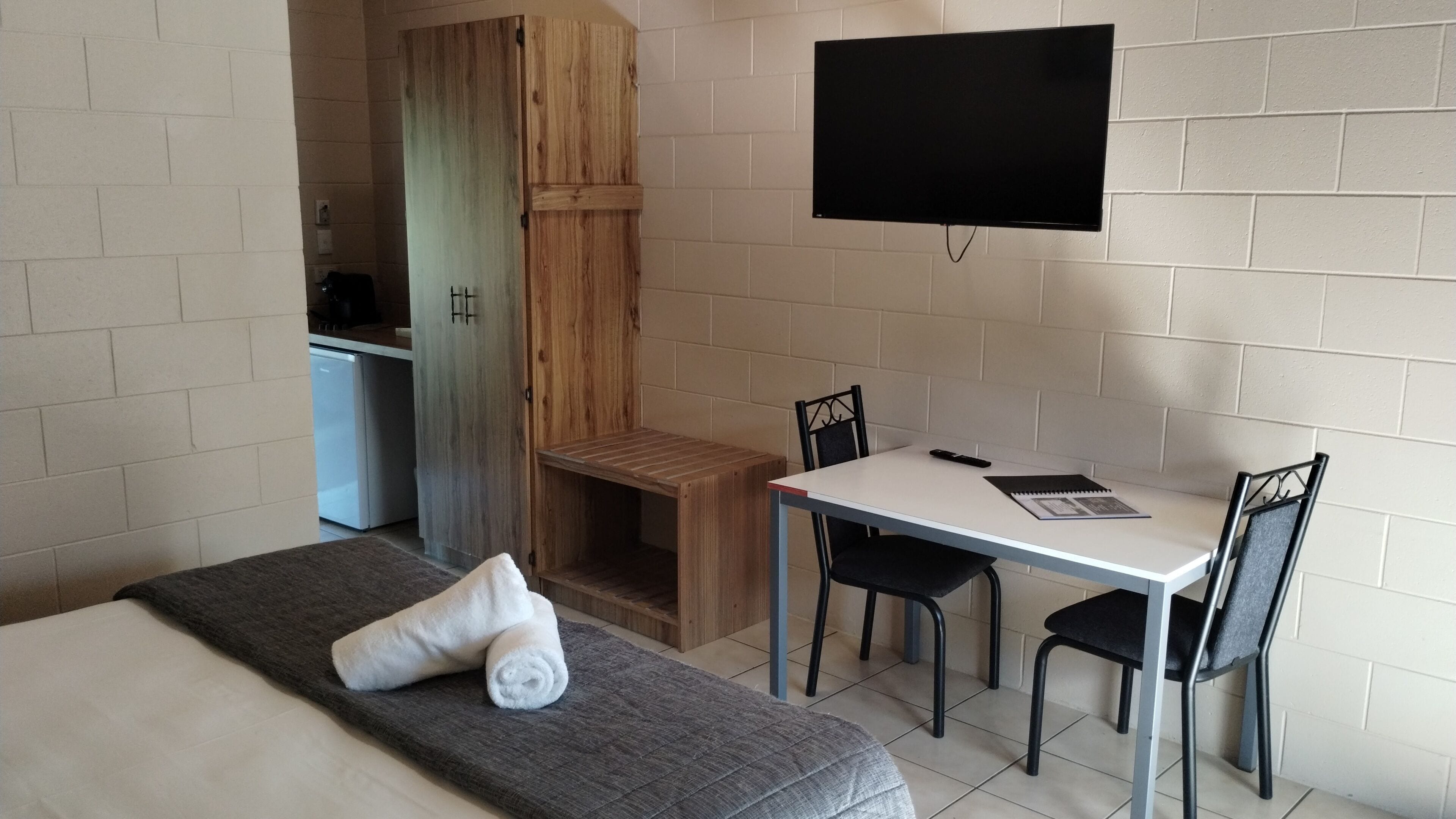 A room with a bed, a desk, two chairs, a TV mounted on the wall, and a small kitchenette in the background.