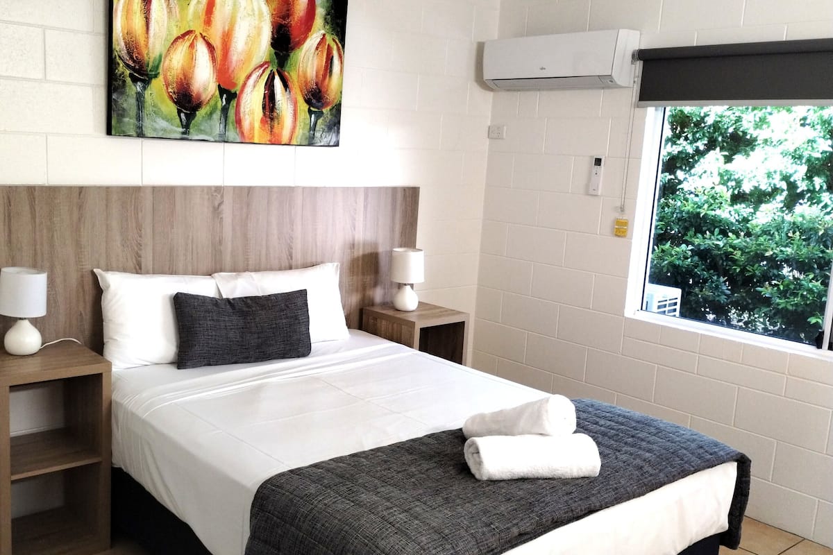 A neatly arranged hotel room with a large bed, two bedside tables with lamps, a painting above the bed, an air conditioning unit, and a window with a view of greenery.