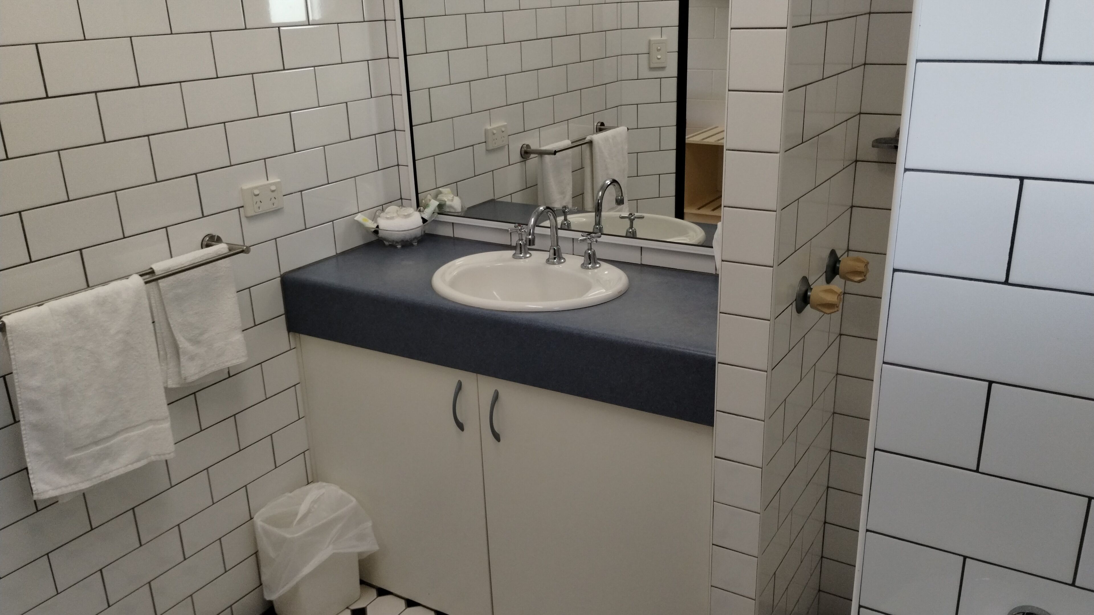 Une salle de bain avec des murs recouverts de carreaux blancs, un lavabo blanc surmonté d’un plan de travail foncé, un miroir au-dessus du lavabo et un porte-serviettes avec une serviette blanche.