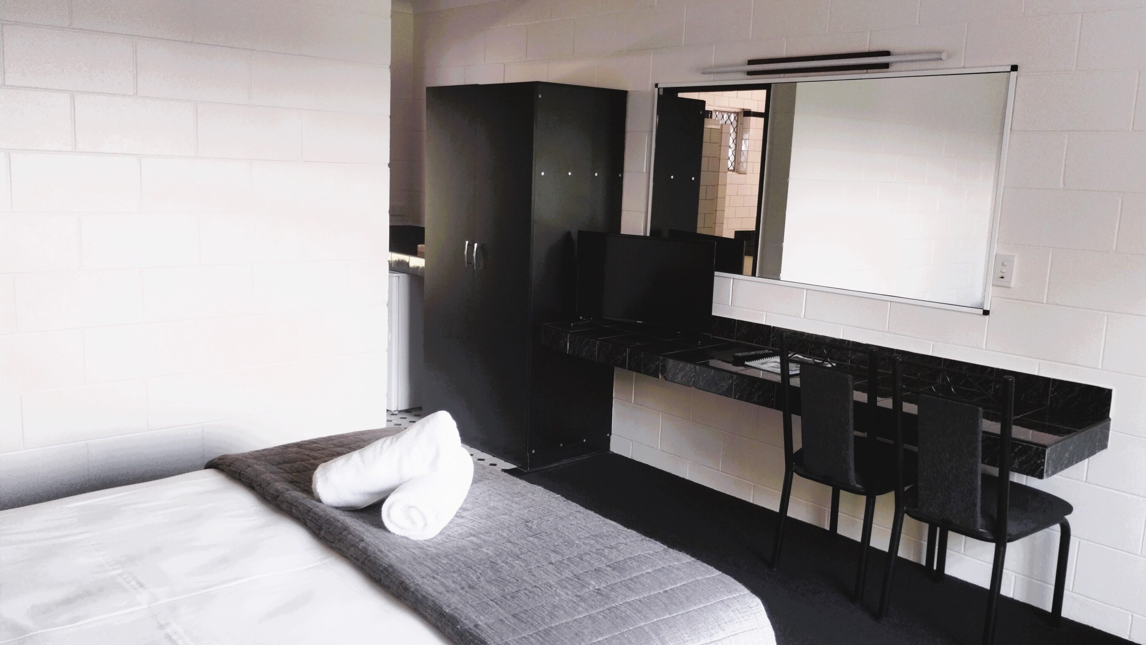 A modern hotel room with a bed, a black desk, a chair, a mirror, and a white tiled wall.