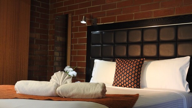 Habitación de hotel con pared de ladrillo, cabecera capitonada, ropa de cama blanca, almohada estampada y un pequeño florero con flores blancas.