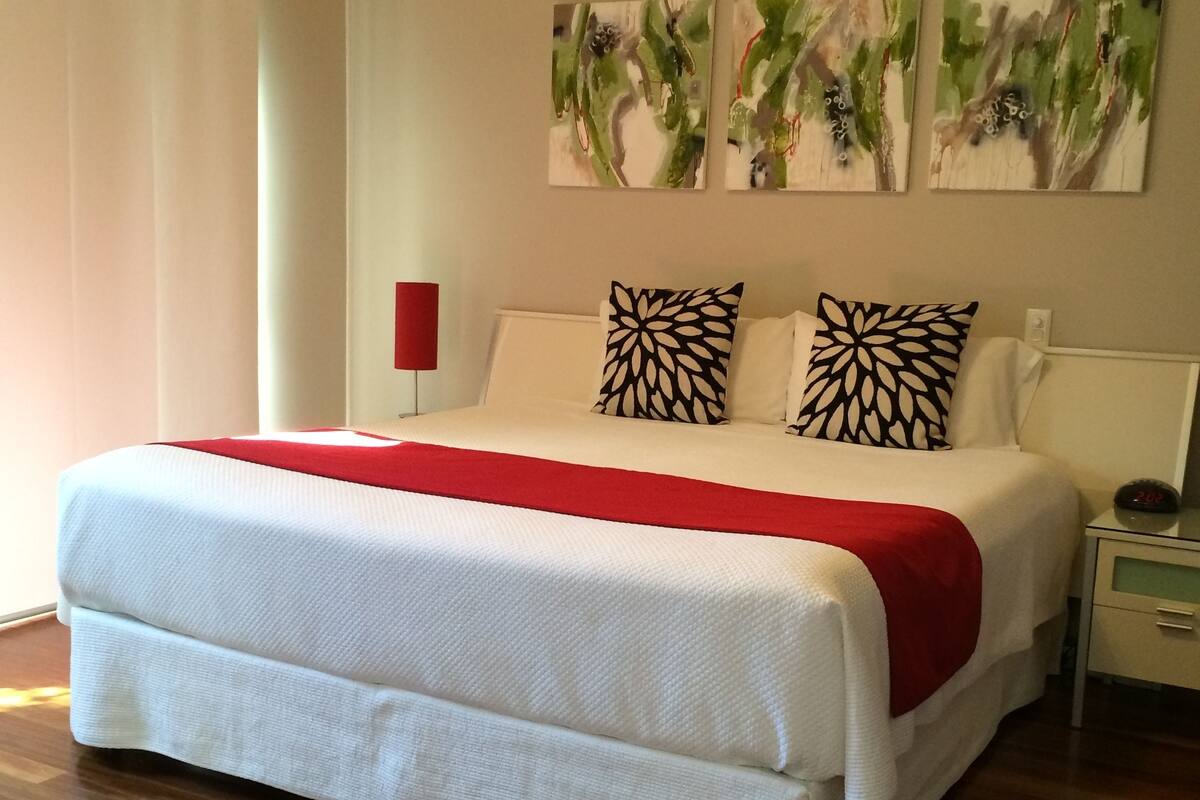 Una cama bien hecha con una colcha roja, almohadas blancas y una lámpara de noche roja. Hay cuadros abstractos en la pared sobre la cama.