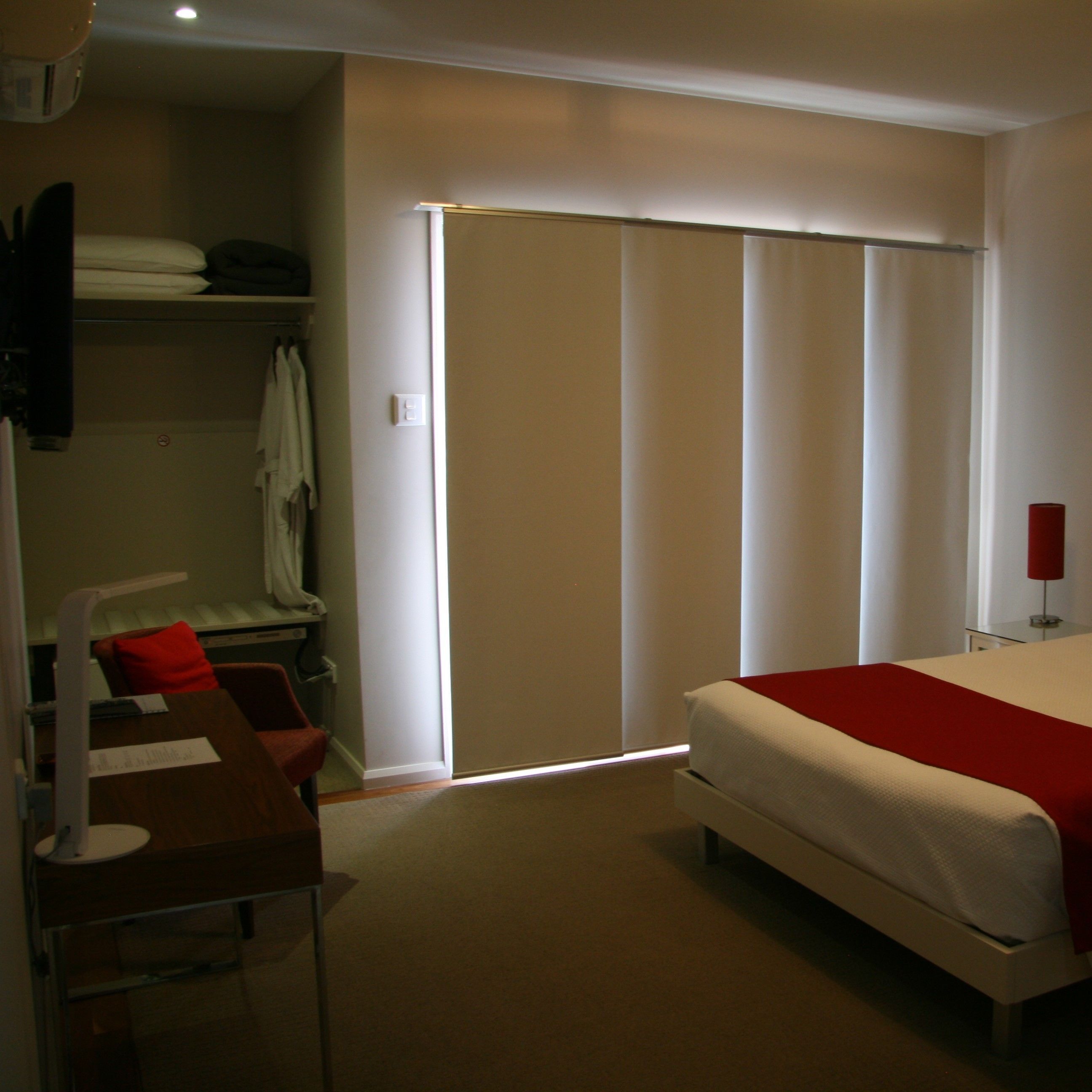 A bedroom with a bed, a desk, a chair, a wardrobe, and a window with blinds.