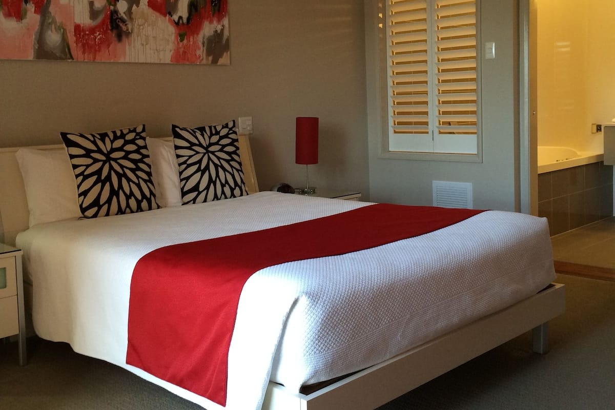 Un dormitorio con una cama grande, dos almohadas, una colcha roja, una mesita de noche, un cuadro en la pared y una ventana con persianas.