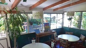 A cozy restaurant interior with wooden beams, a large window offering a view of the outdoors, and a variety of seating options including a round table and chairs.