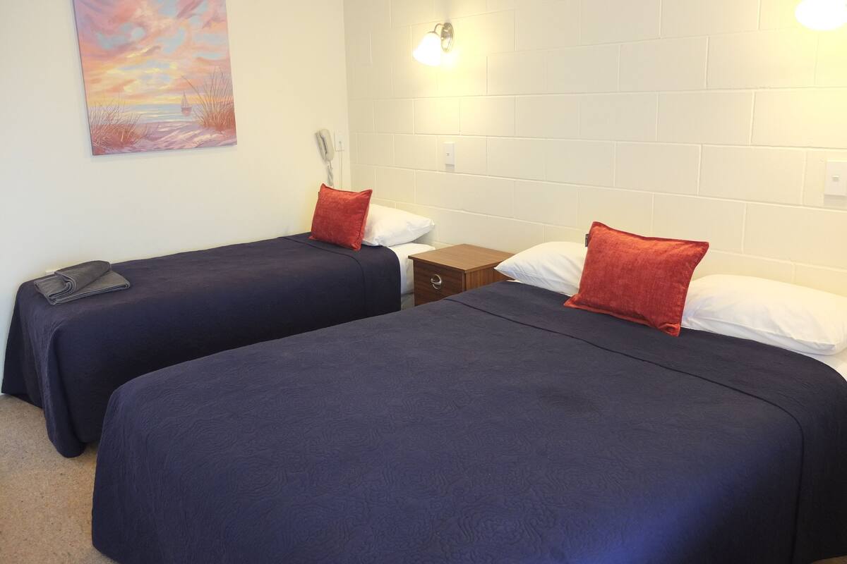 A room with two beds, each with a dark blue bedspread and a red pillow. There is a painting on the wall and a white brick wall.