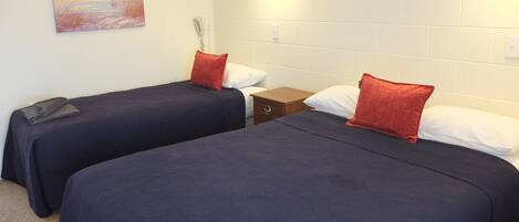 Una habitación con dos camas, cada una con una colcha azul oscuro y una almohada roja. Hay un cuadro en la pared y una pared de ladrillo blanco.