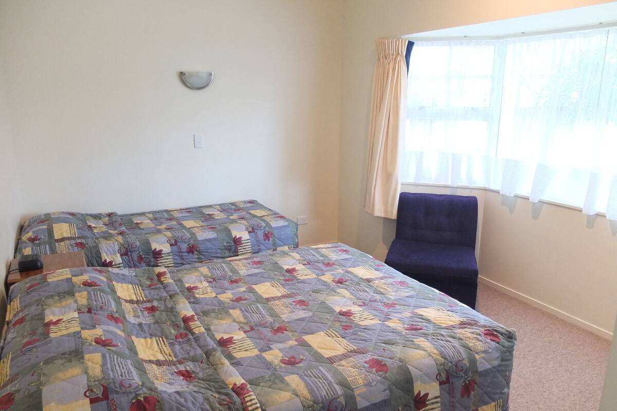 A room with two beds covered in a patchwork quilt, a chair, a wall-mounted light fixture, and a window with curtains.