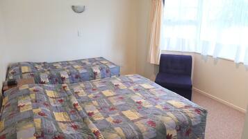 Habitación con dos camas cubiertas con un edredón de retazos, una silla, una lámpara fijada a la pared y una ventana con cortinas.