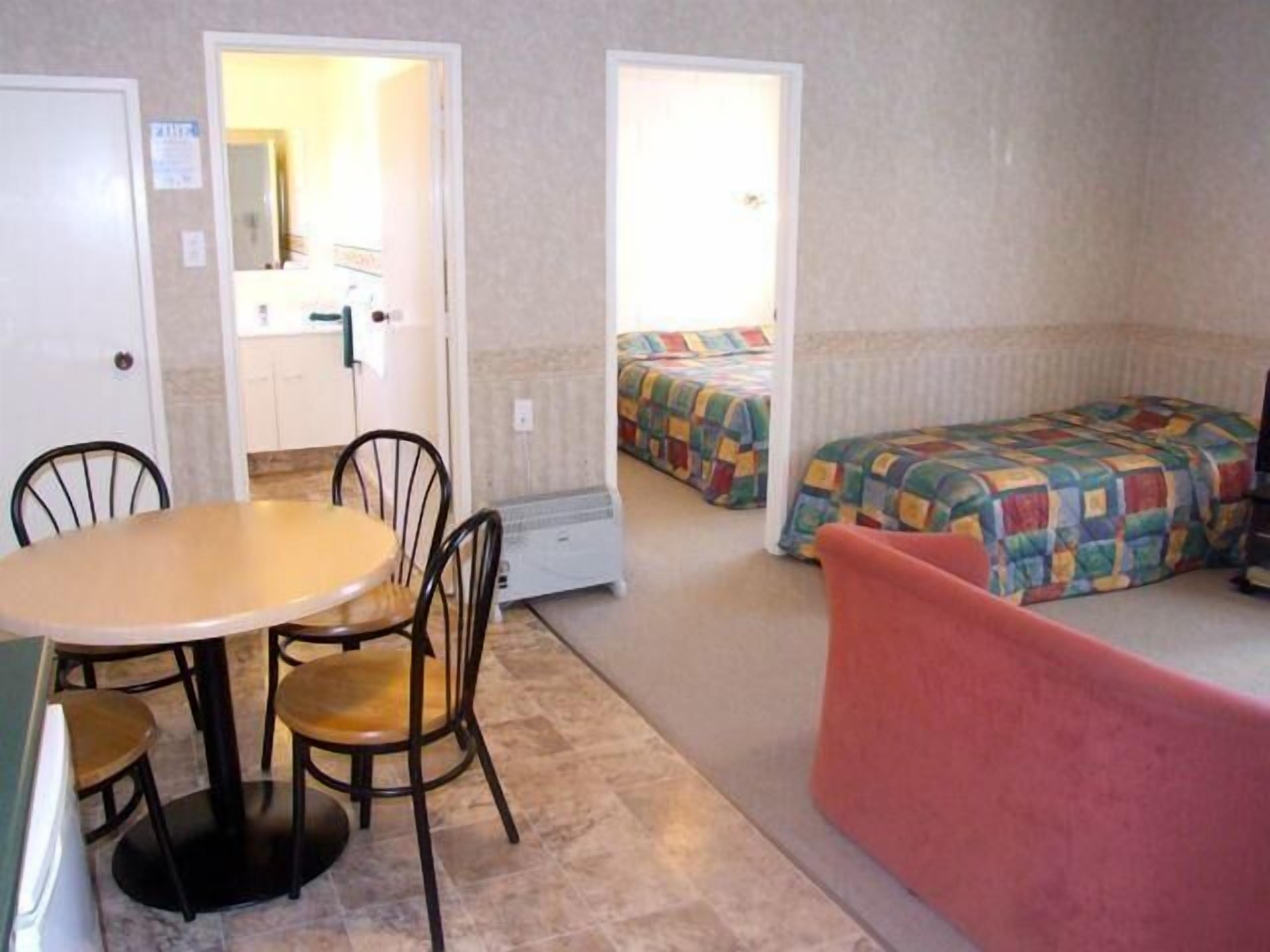 A compact room with a round table and four chairs, a bed with a colorful quilt, and a red couch.