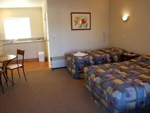 A room with two beds, a small table, and a chair. There is a kitchenette area with a sink and a window.