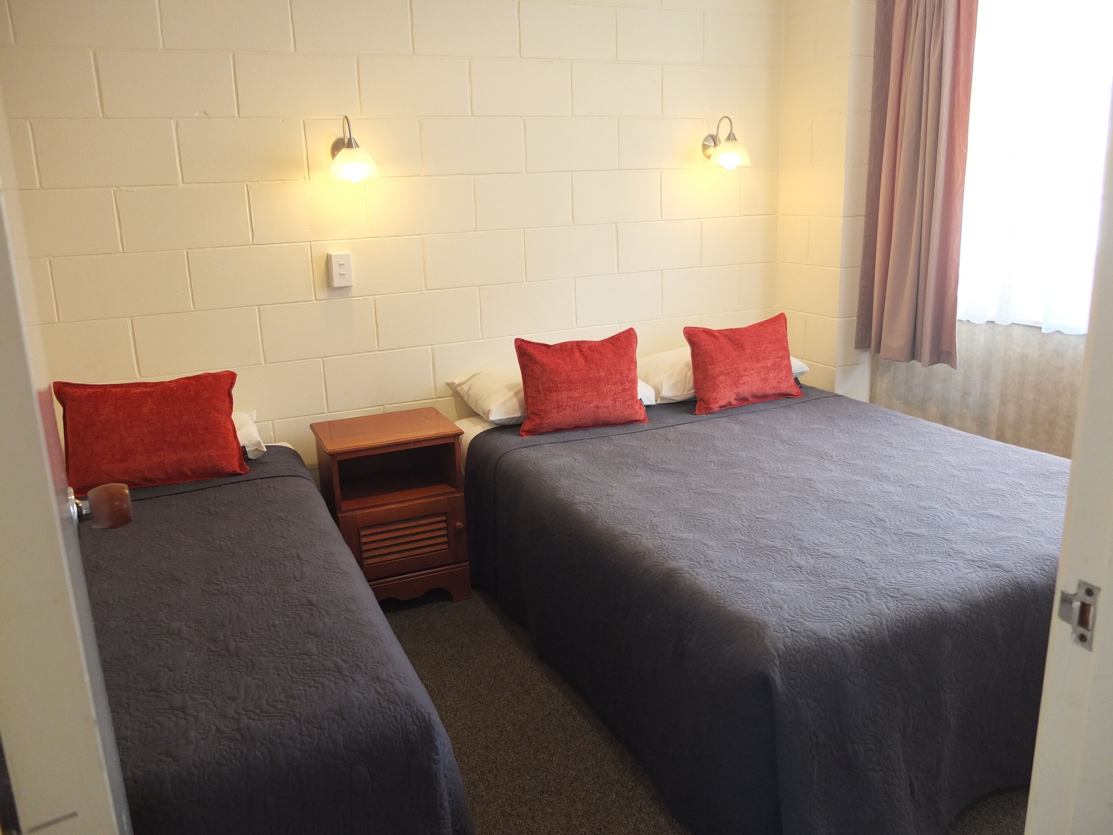 Une chambre double avec deux lits, chacun agrémenté d’un oreiller rouge, d’une table de chevet en bois et de deux lampes fixées au mur.