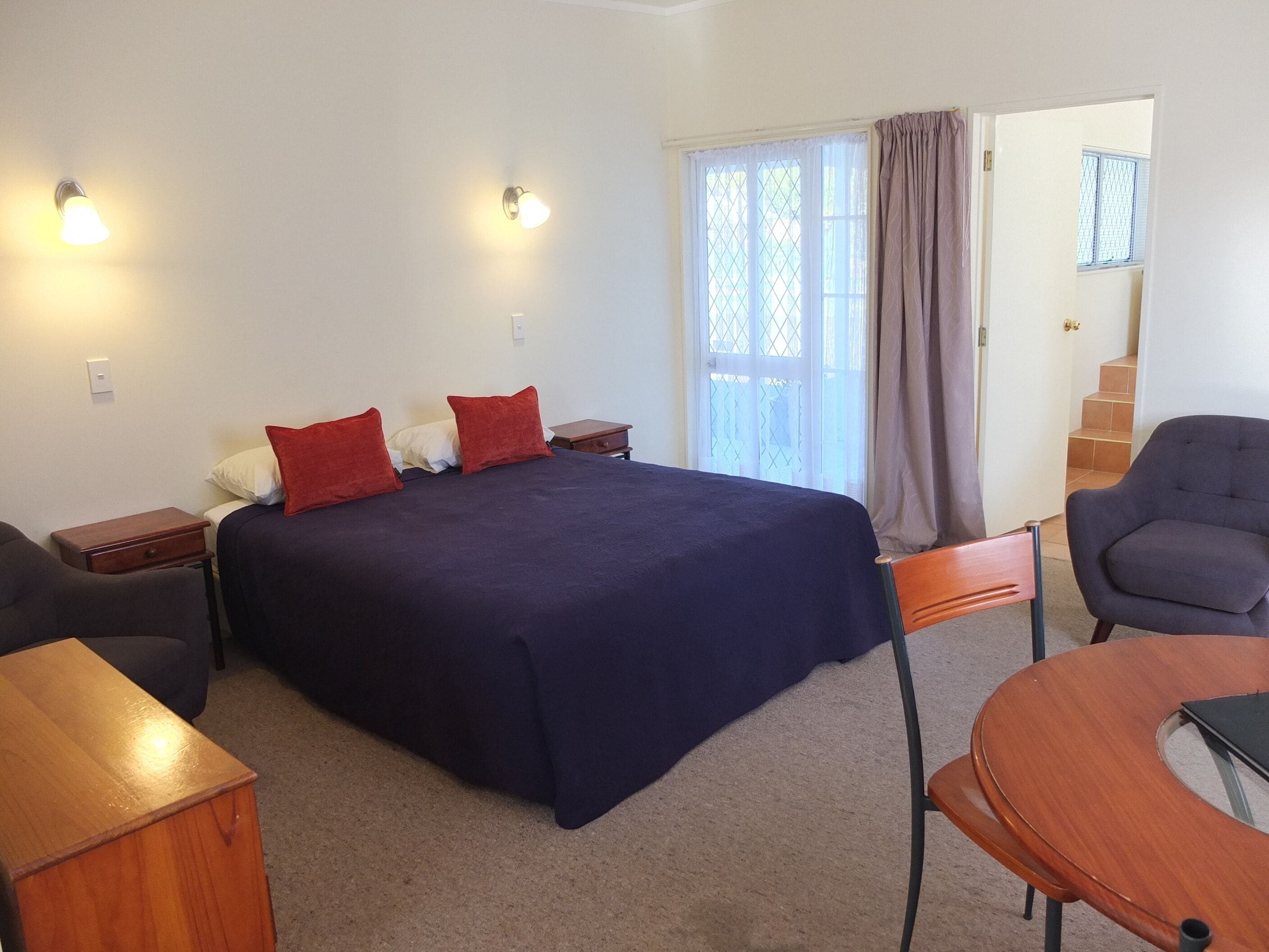 A bedroom with a bed, two bedside tables, a chair, a wooden side table, a window with curtains, and a door leading to a staircase.