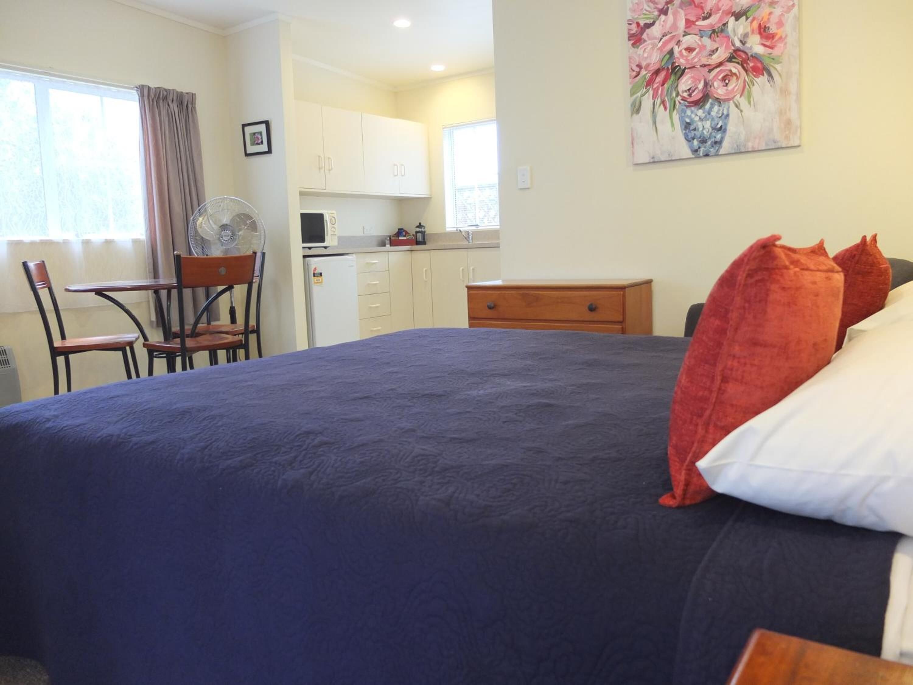 A bedroom with a large bed, a chair, a desk, and a kitchenette in the background.