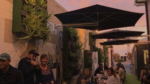 Un espace extérieur aménagé pour les repas, avec des tables et des parasols, entouré de verdure et bordé par la façade d’un bâtiment ornée de graffitis.