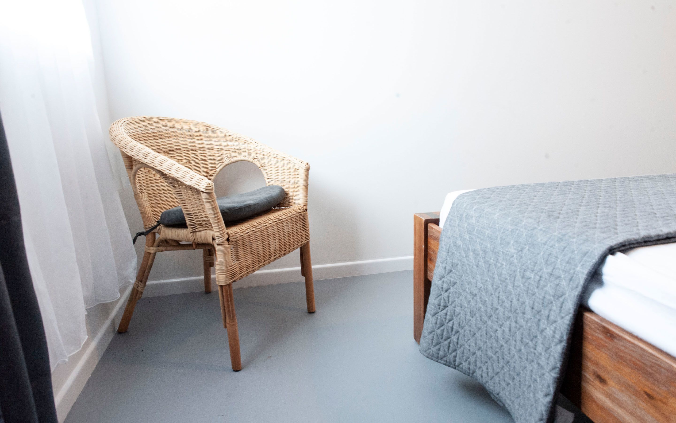 Una habitación con una silla de mimbre, una cama con colcha gris y una pared blanca.
