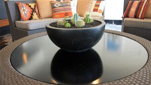 Une table ronde avec un plateau noir et un bord tressé, entourée d’une chaise également tressée. Au fond, on aperçoit un canapé agrémenté de coussins colorés.