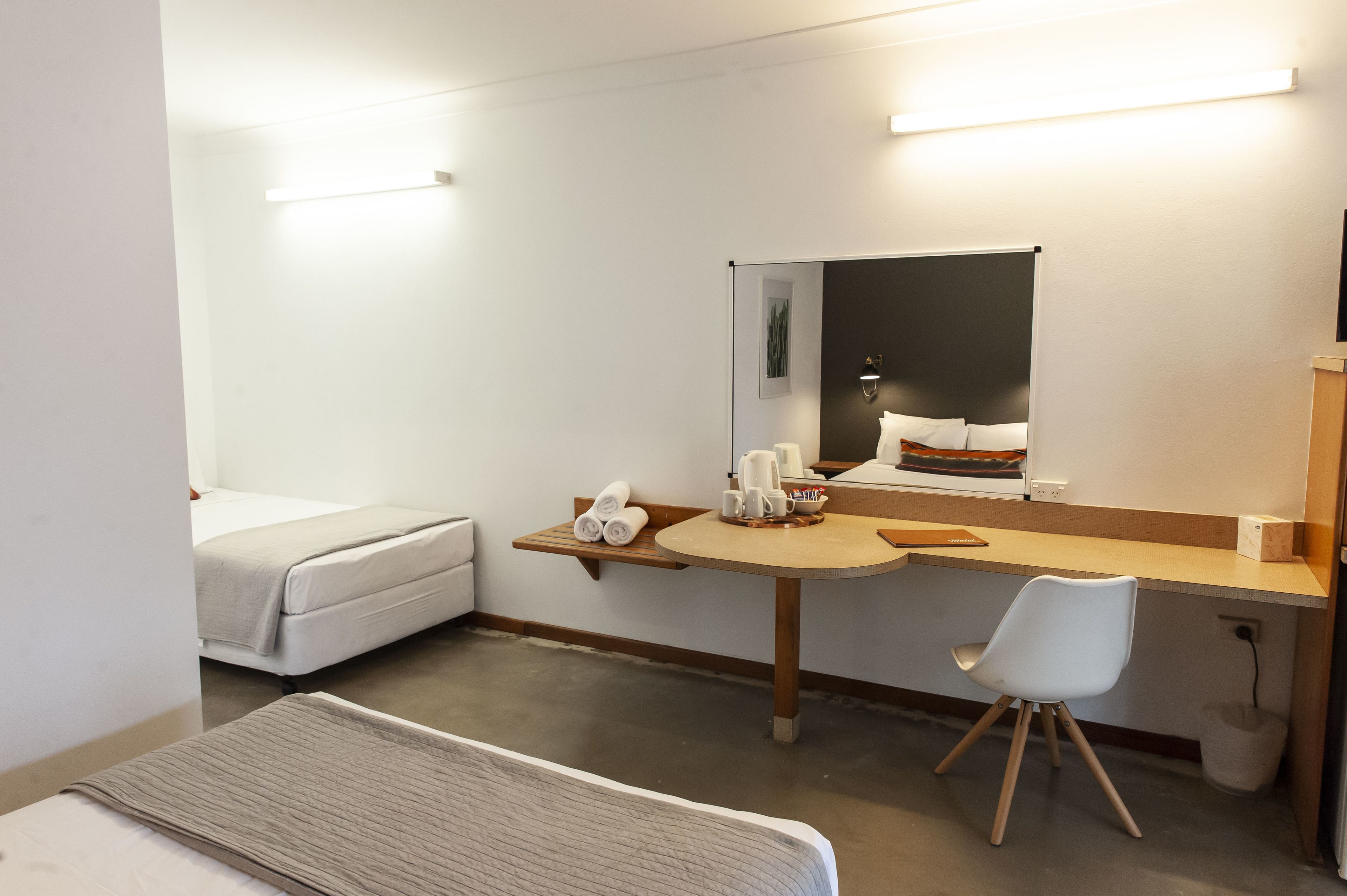 A hotel room with a bed, a desk with a chair, a mirror, and a small table with towels and mugs.