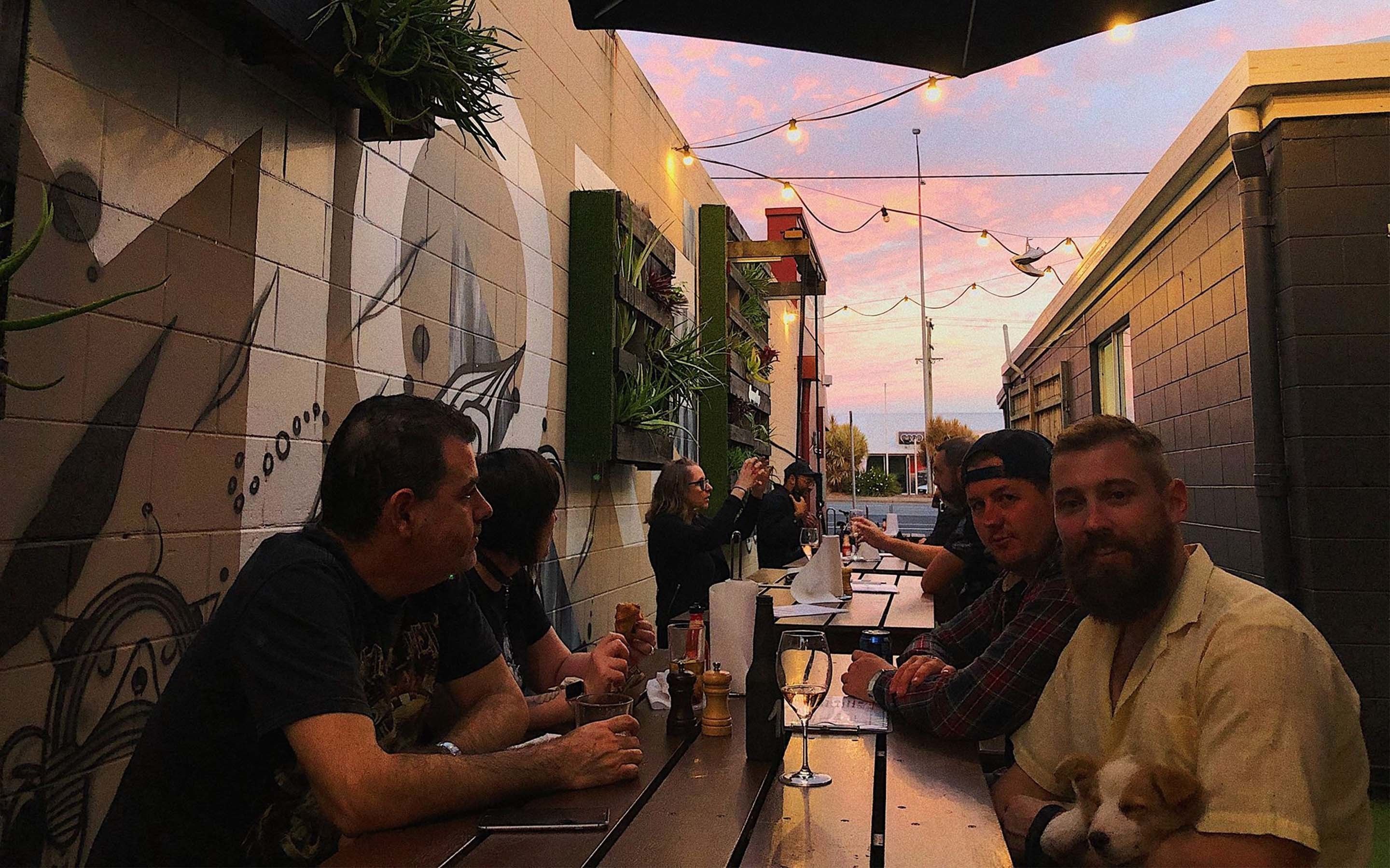A group of people dining at an outdoor restaurant with a tiled wall and plants on the side.