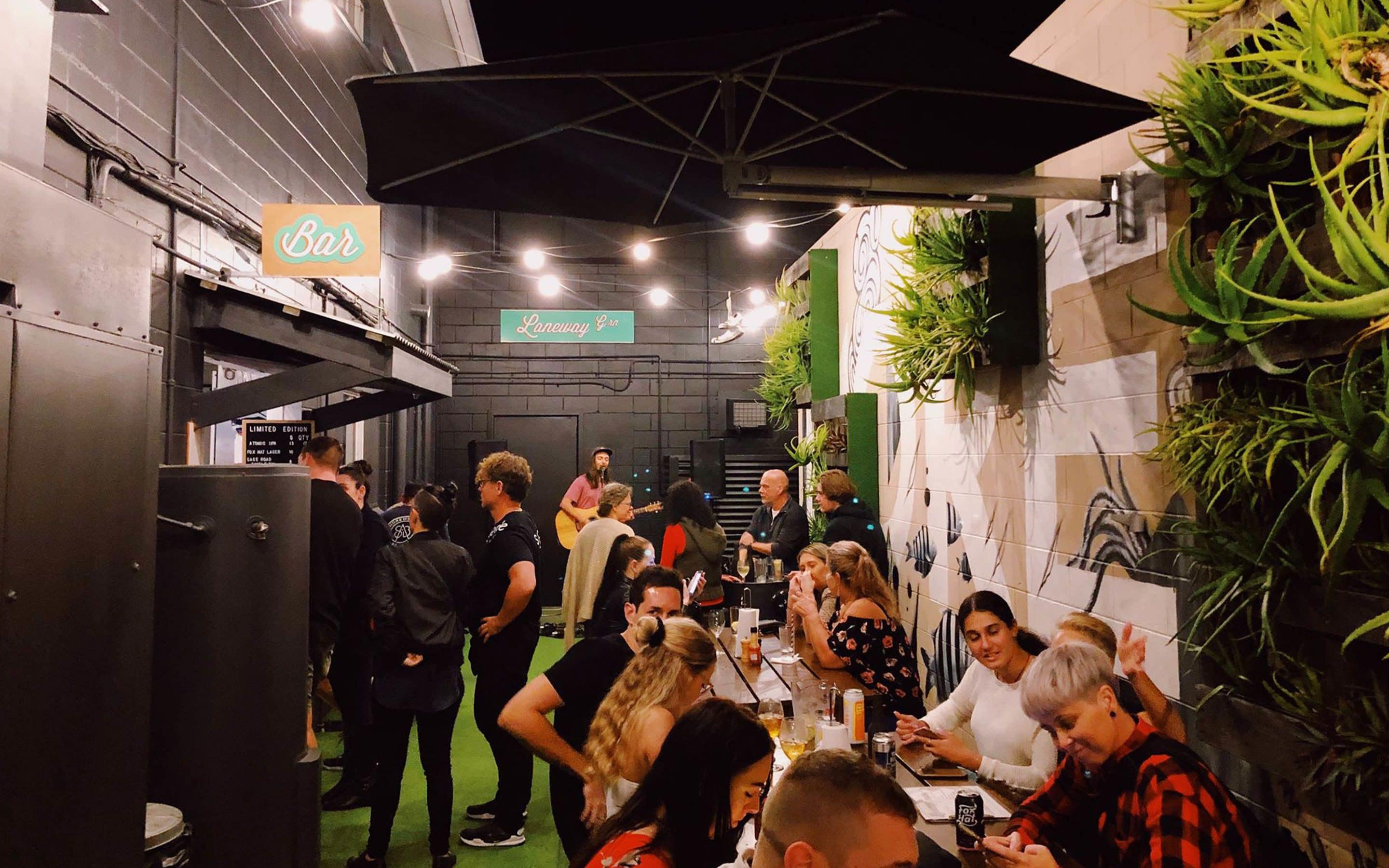 Zona de bar con gente conversando, un letrero que dice 'Laneway Grill' y un cartel que pone 'Bar' sobre la entrada.