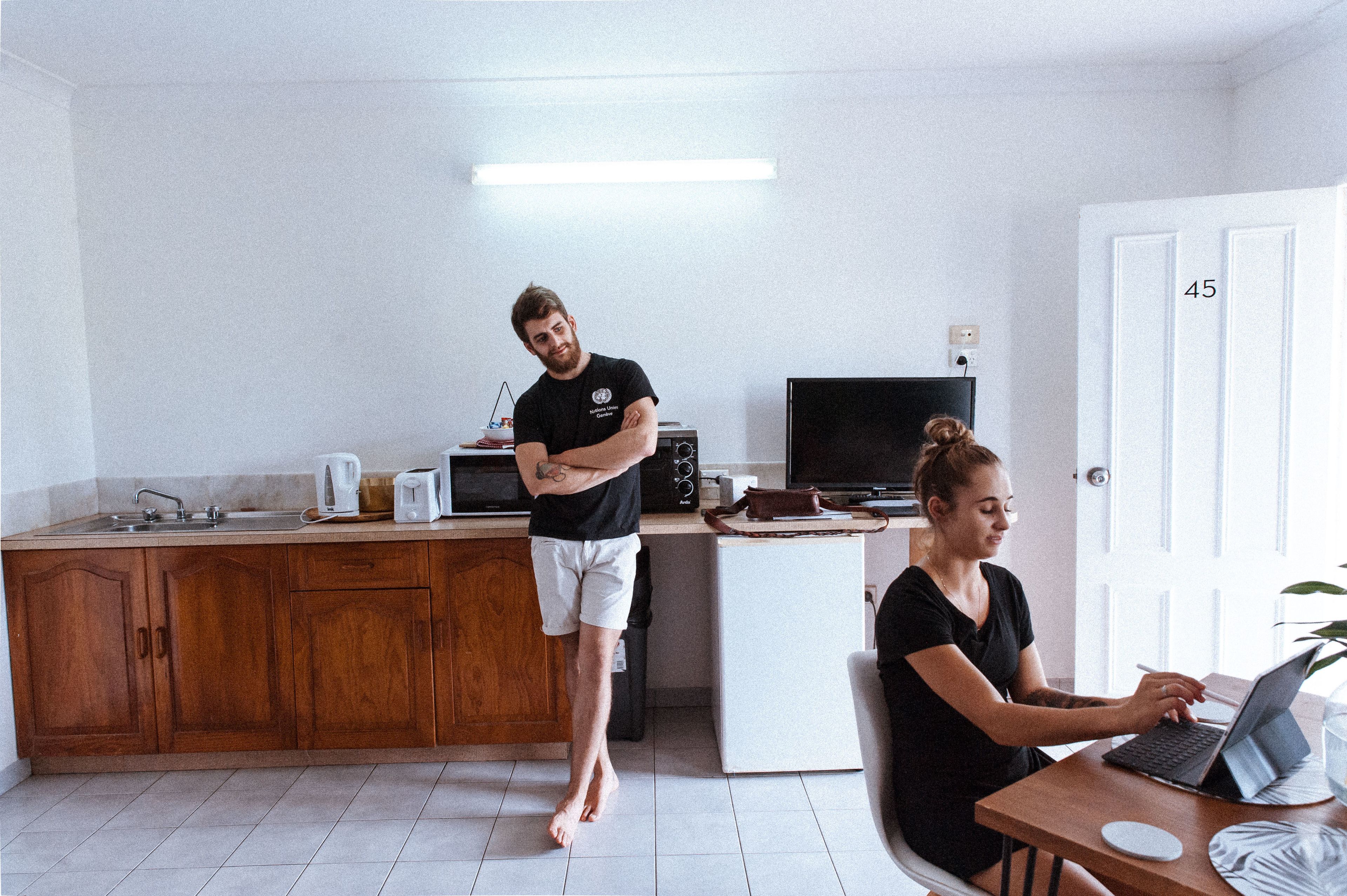 A small, modern kitchen with wooden cabinets, a microwave, and a refrigerator. A person is standing with arms crossed, and another is sitting at a table using a laptop.