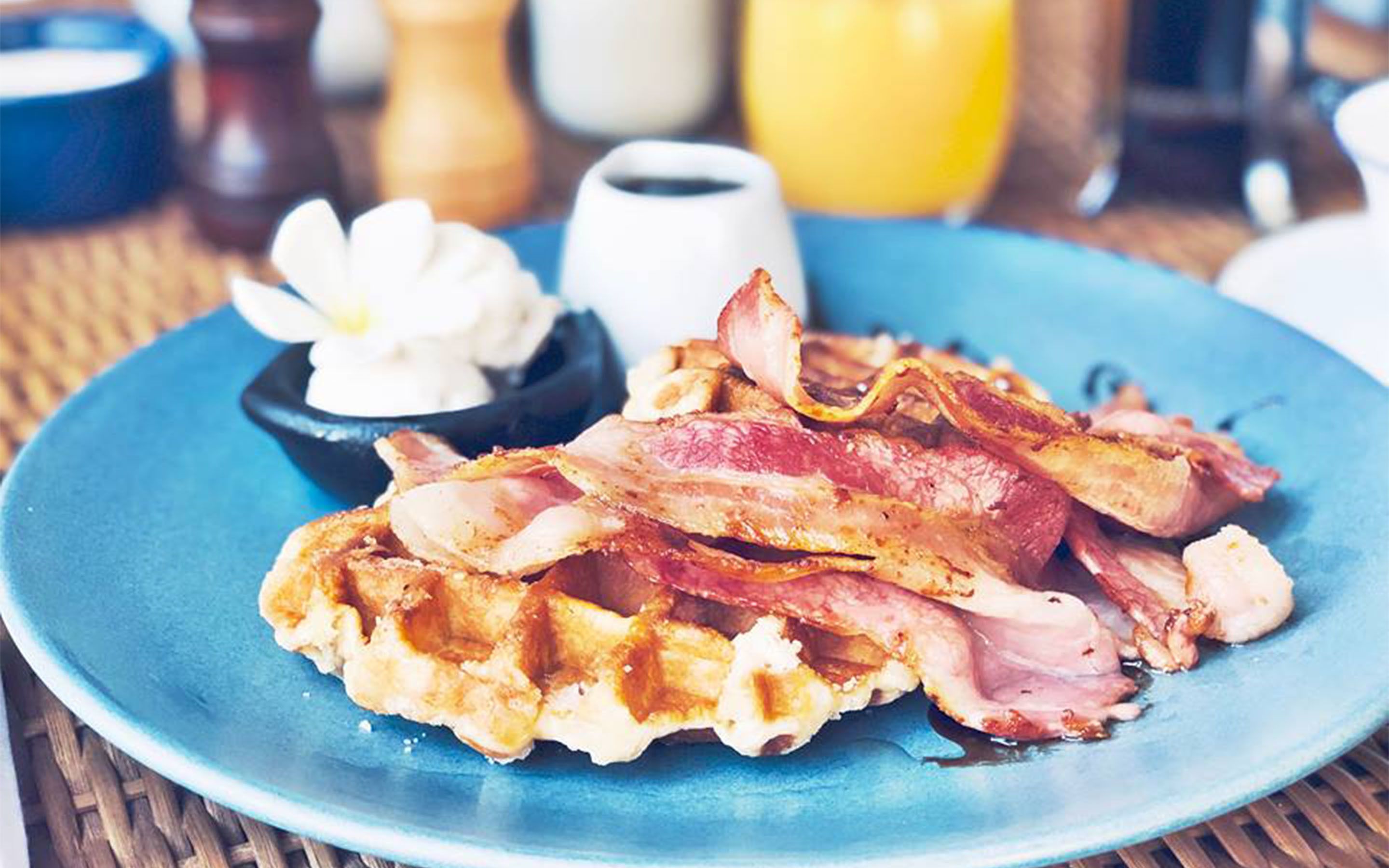 Bacon und Waffeln auf einem blauen Teller, im Hintergrund eine Blume und Beilagen.