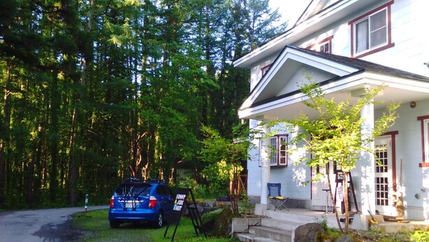 A house with a covered porch, a car parked in front, and a forested area in the background. - Sunny Side Hut (Hakuba)