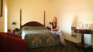 A hotel room with a bed, a chair, a nightstand, and a table with a lamp and a coffee maker. - Lucas Hotel (Latrobe)