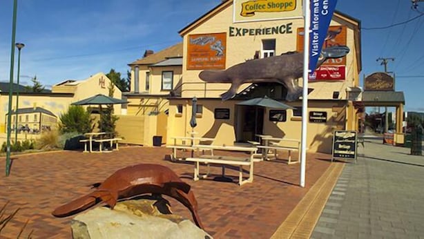 A building with a sign for 'Coffee Shopper' and 'Experience'. There are picnic tables and a dinosaur sculpture in the foreground. - Lucas Hotel (Latrobe)