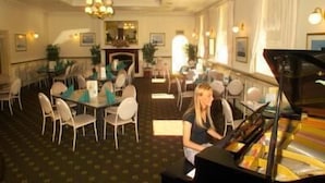 A hotel dining area with round tables, chairs, a piano, and a fireplace. - Lucas Hotel (Latrobe)