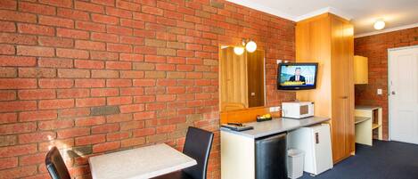 Una habitación pequeña con pared de ladrillo, un escritorio con silla, televisión, microondas y refrigerador compacto.