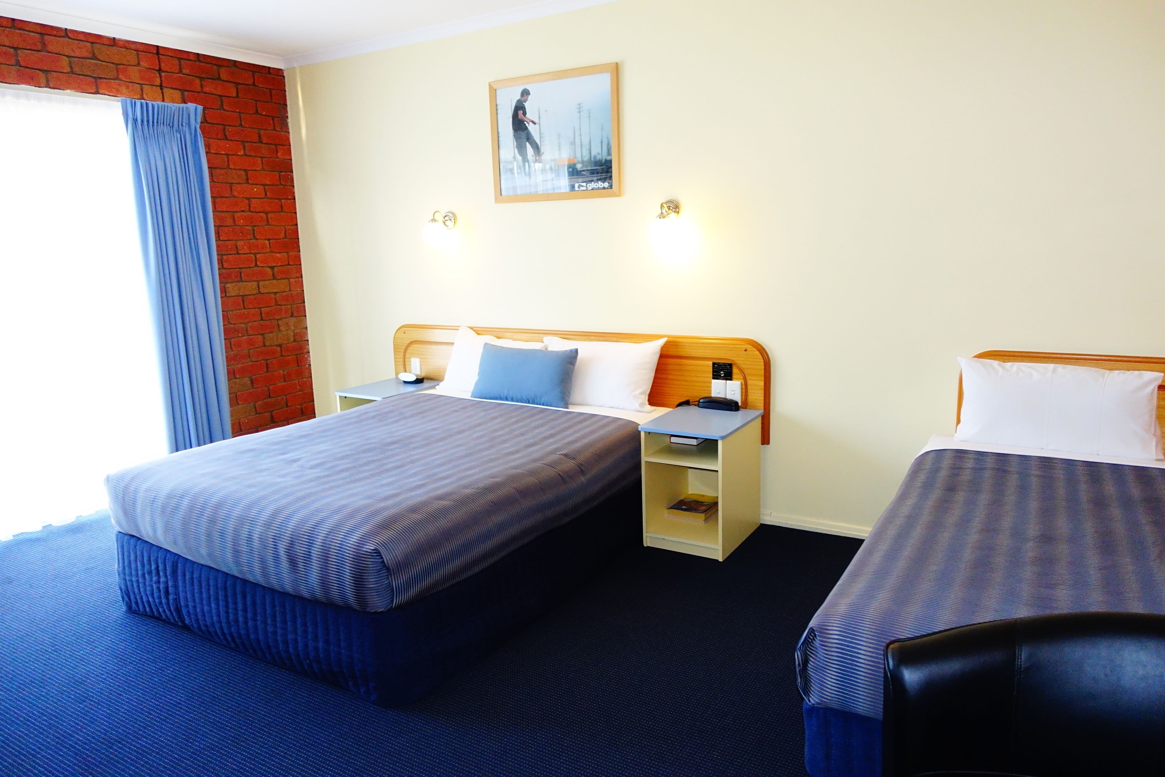 A hotel room with two beds, a brick wall, a window with blue curtains, and a painting on the wall.