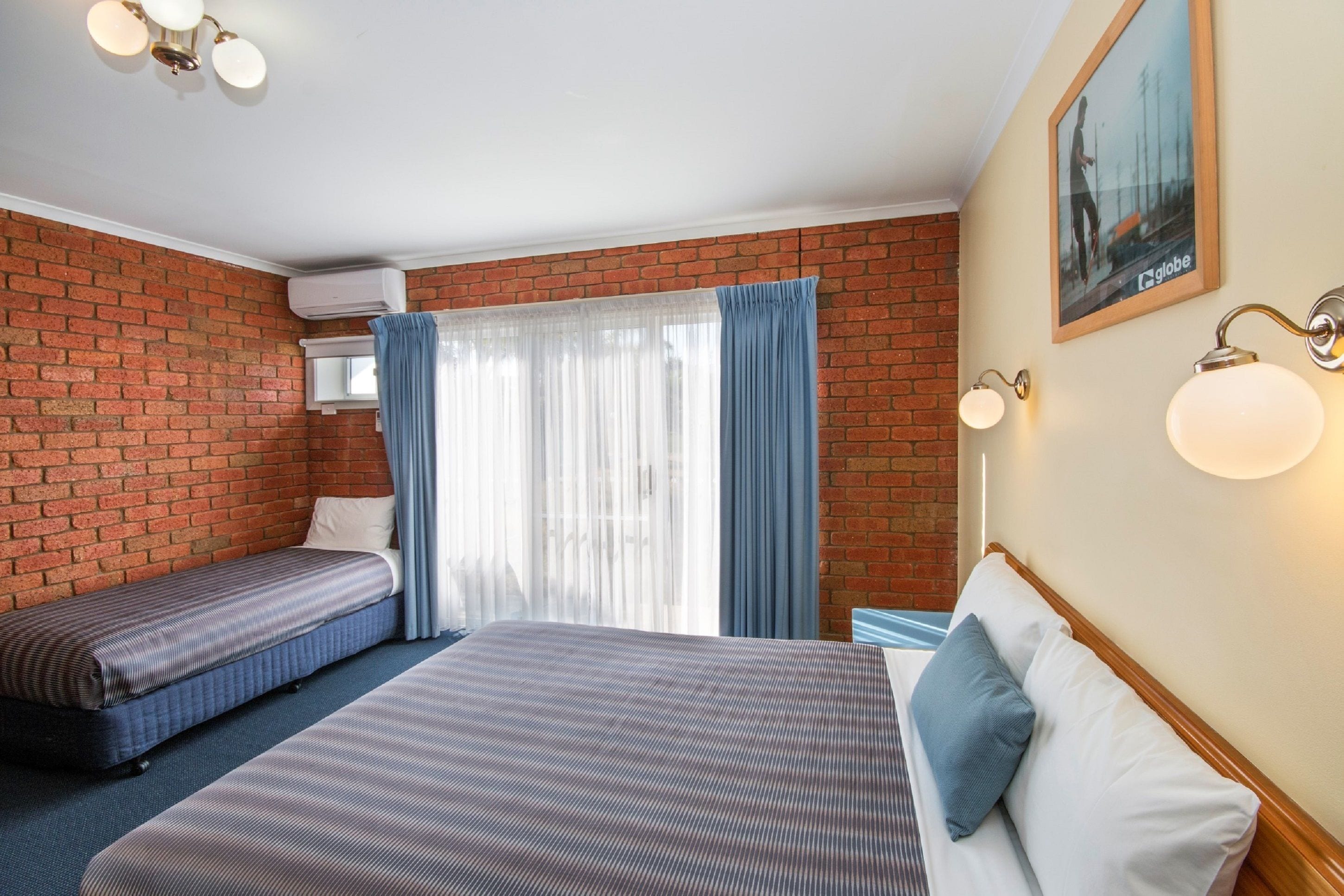 A hotel room with a brick wall, a large bed with a blue pillow, a smaller bed, a window with sheer curtains, and a wall-mounted air conditioning unit.